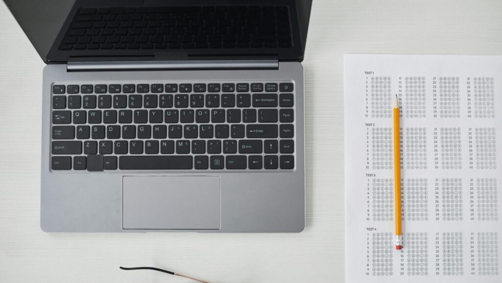 Flat lay of a laptop, exam sheet, and pencil on a white desk, perfect for education and technology themes.