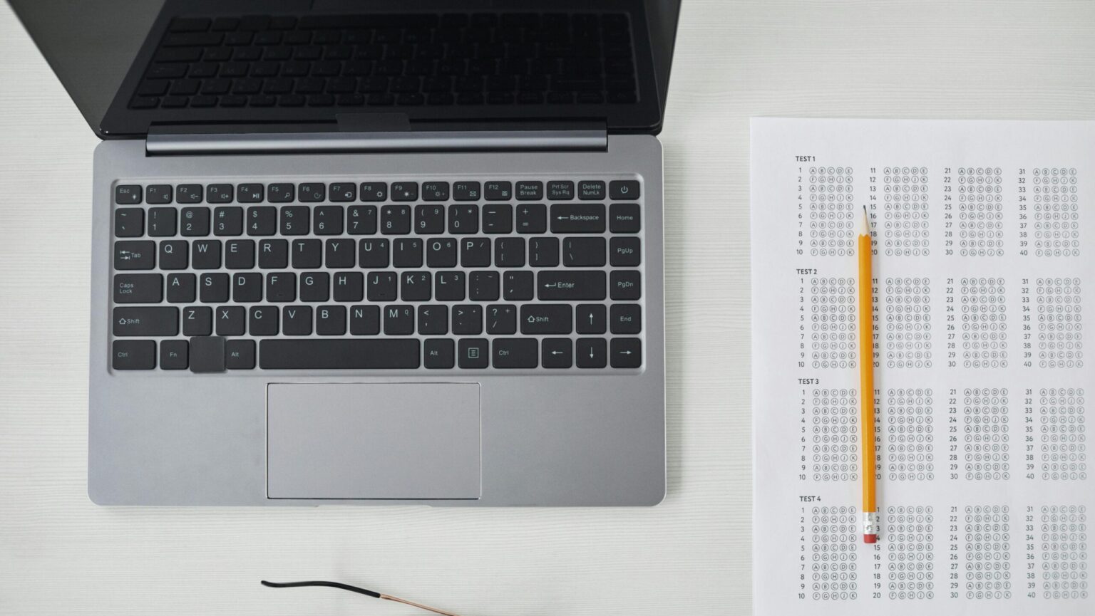 Flat lay of a laptop, exam sheet, and pencil on a white desk, perfect for education and technology themes.