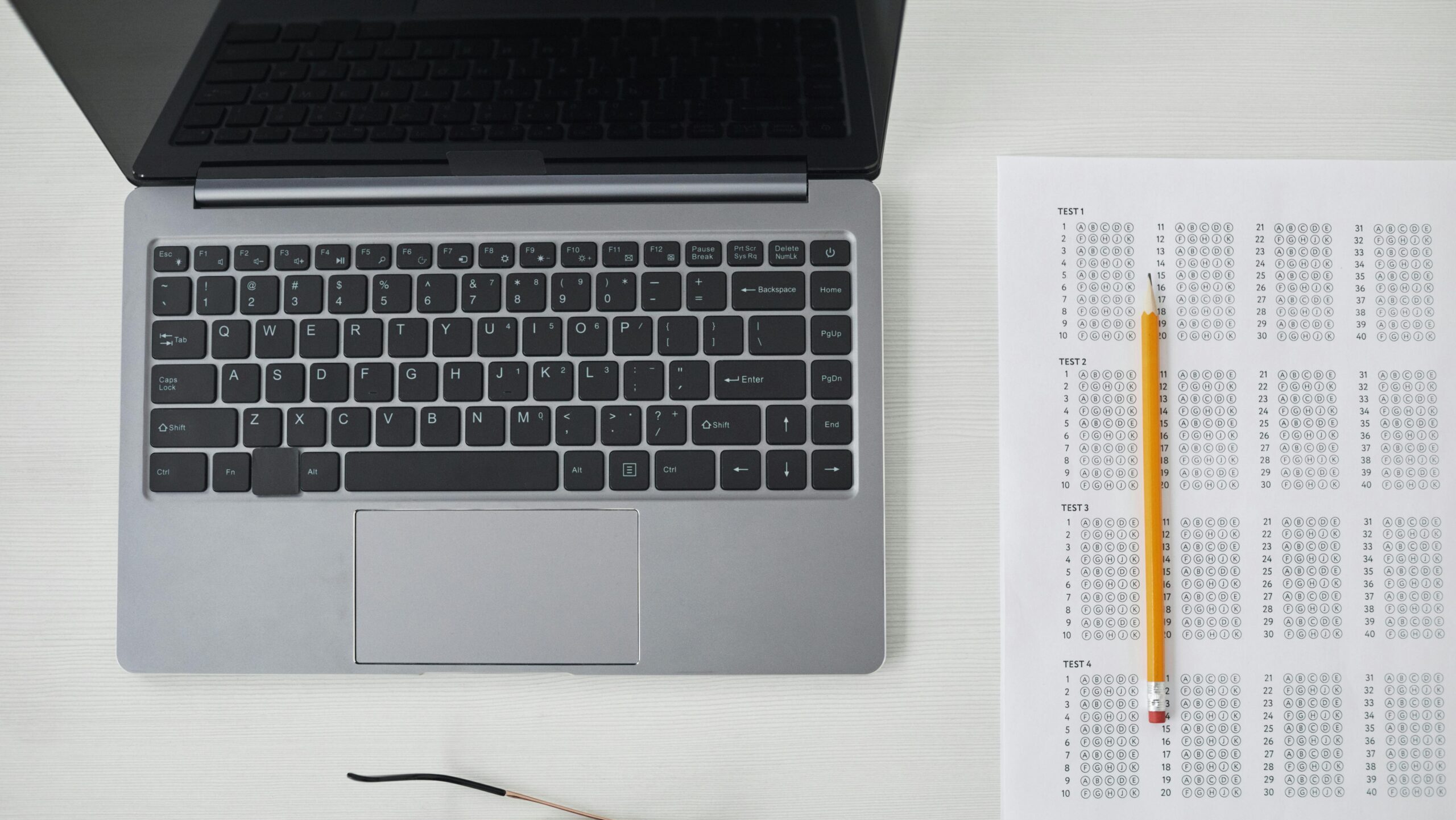 Flat lay of a laptop, exam sheet, and pencil on a white desk, perfect for education and technology themes.