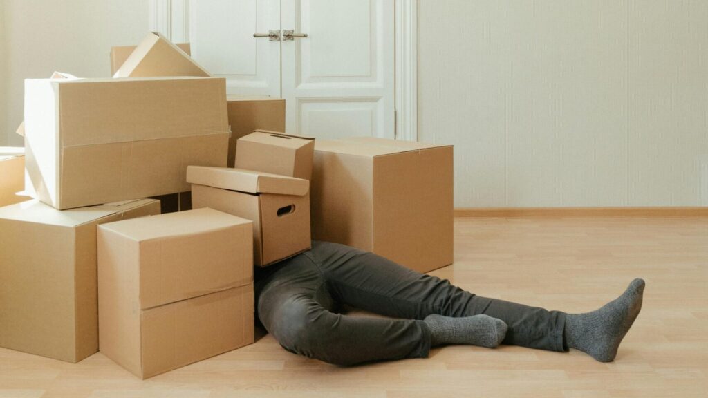 A person lying under a pile of moving boxes in a minimalistic room conveys tiredness from packing.