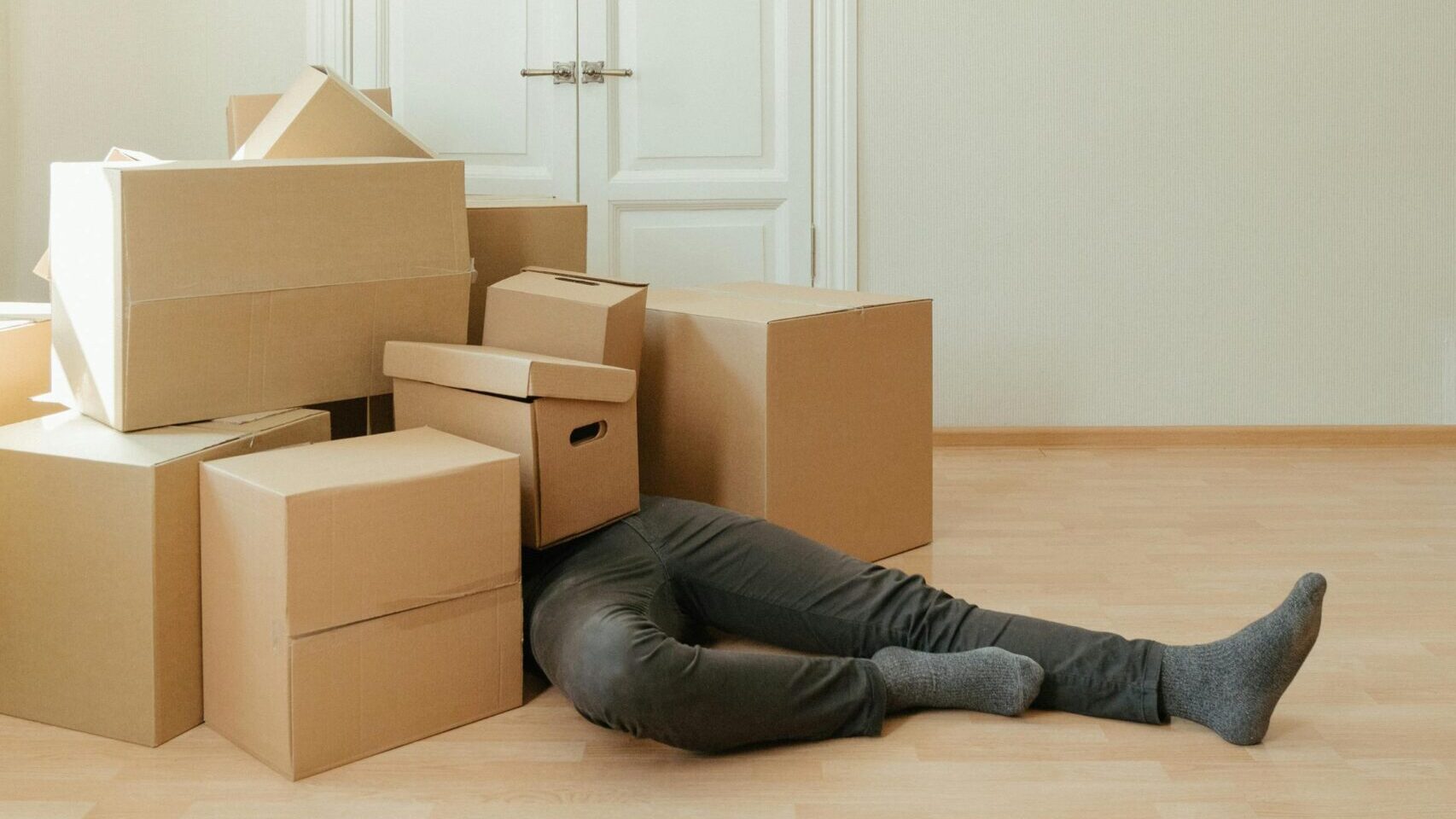 A person lying under a pile of moving boxes in a minimalistic room conveys tiredness from packing.